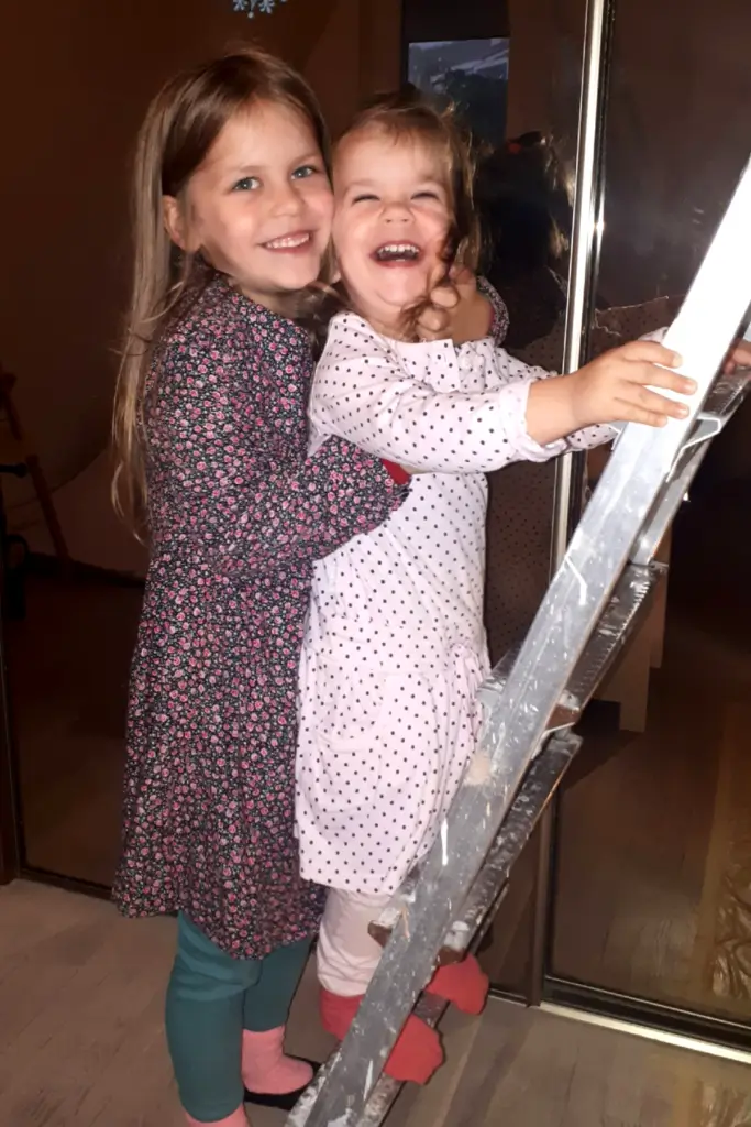 How to Paint Baby Room Wall 14 Two smiling young girls hugging by a ladder indoors, cozy home moment.
