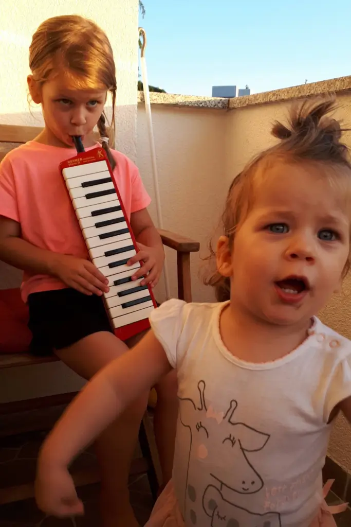 How to Deal with Toddlers Fighting Over Toys 3 Two young kids on a balcony, toddler dancing while sister plays a red melodica.