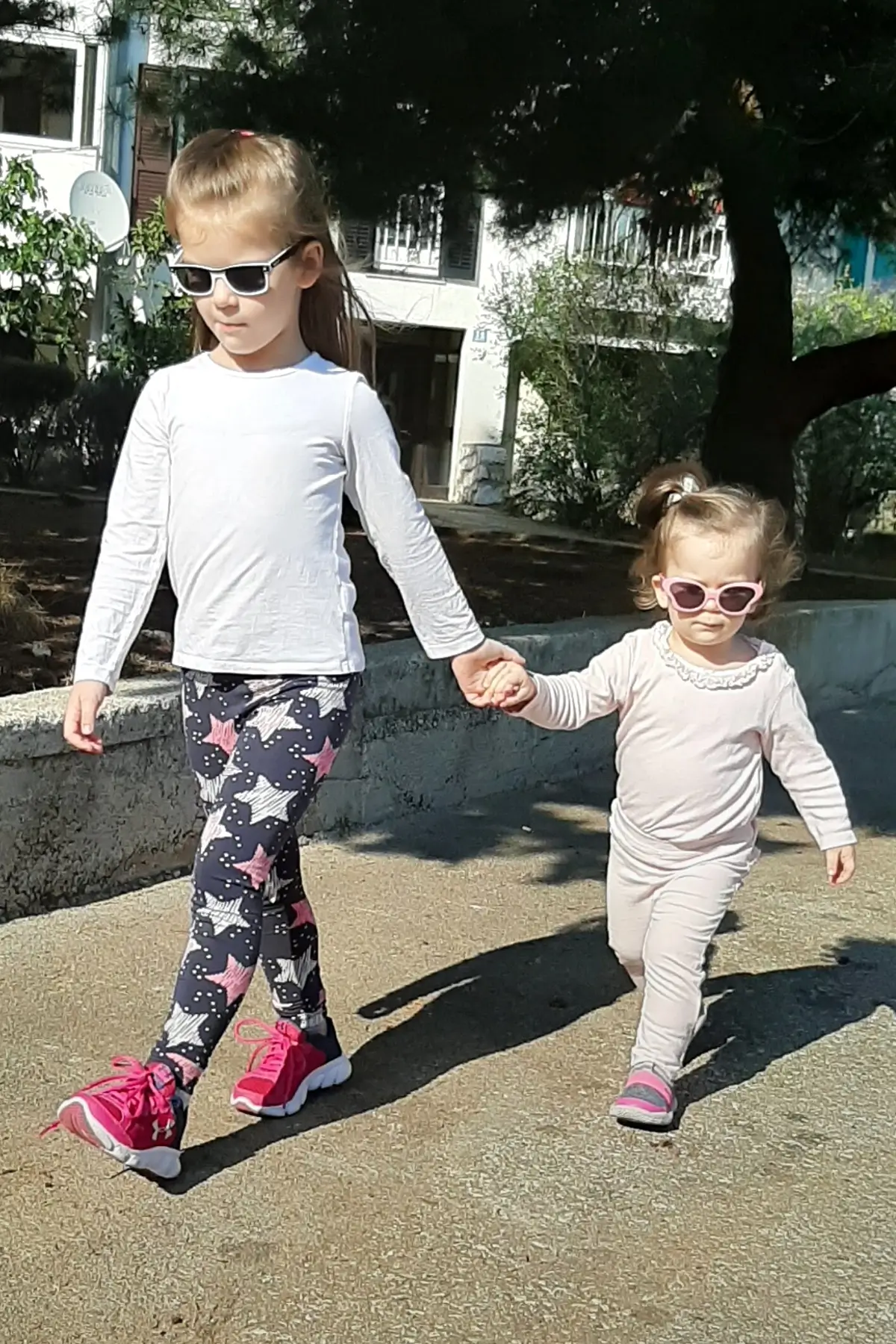 How to Deal with Toddlers Fighting Over Toys 7 Two young sisters wearing sunglasses hold hands while walking outside on a sunny day.