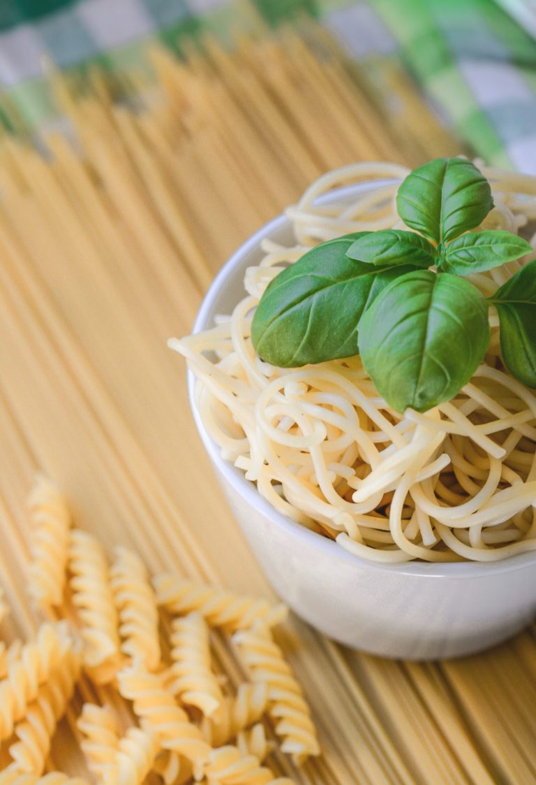 Creamy Pesto Pasta: A Harmony of Freshness and Indulgence 3 Bowl of cooked spaghetti with fresh basil, surrounded by raw spaghetti and fusilli pasta on a kitchen table.
