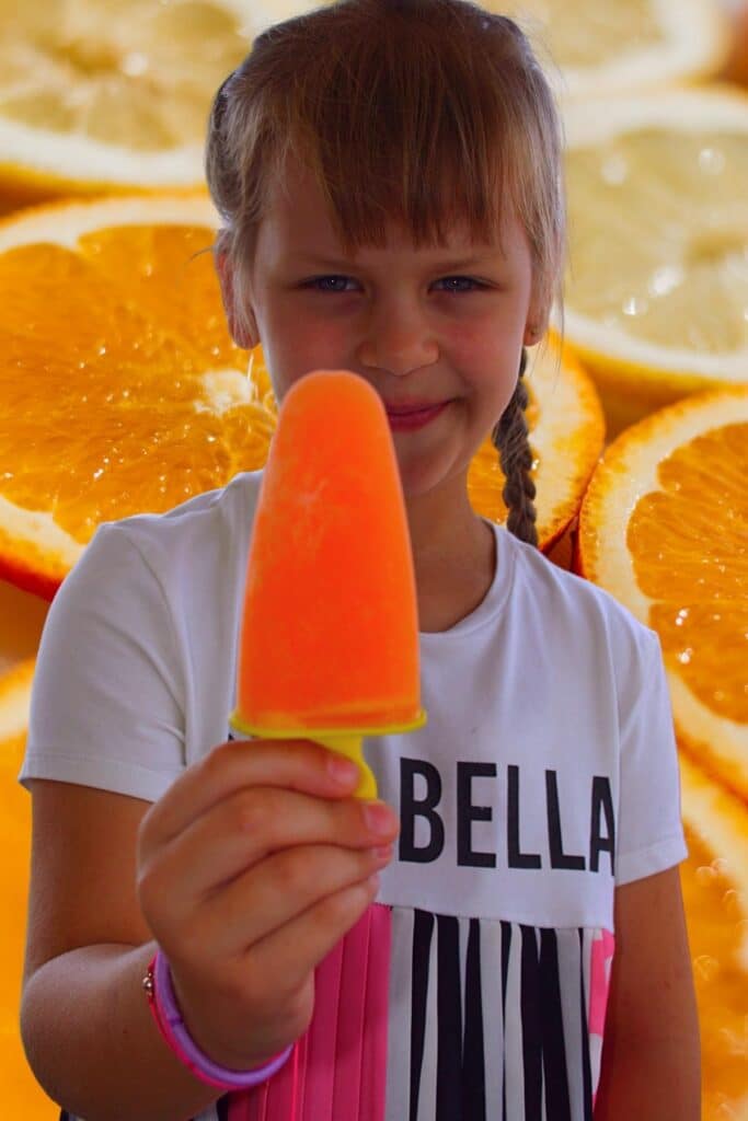 Child holding homemade orange popsicle with fresh orange slices background