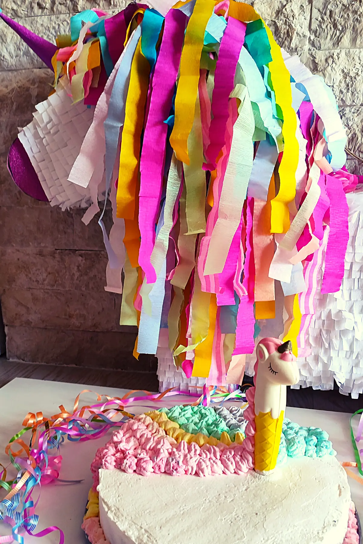 Unicorn piñata with rainbow streamers above unicorn-themed birthday cake and ribbons decoration.