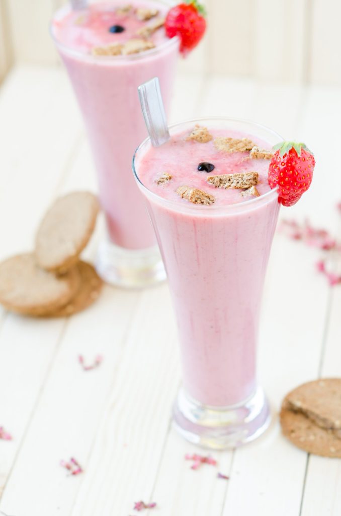 Tall glasses of strawberry smoothie with granola crumble and fresh berries on a light wooden table
