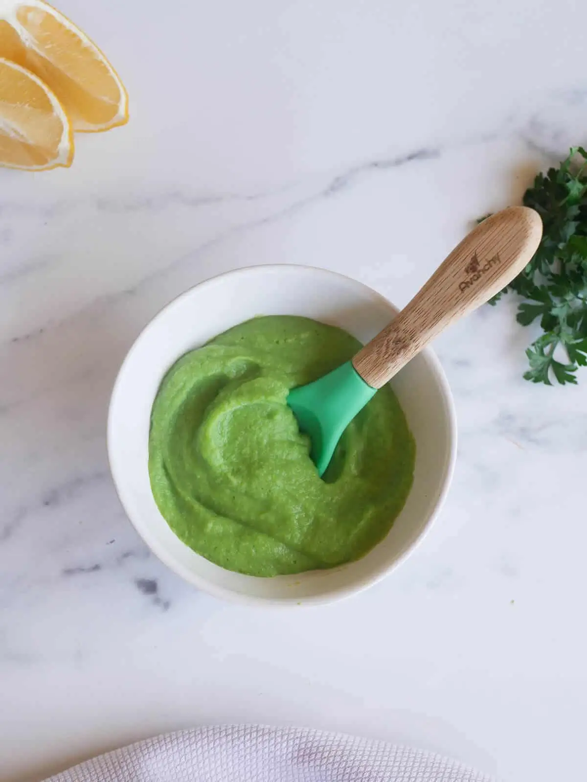 Homemade pea puree in white bowl with baby spoon, lemon and parsley